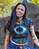 Woman wearing a tie-dye shirt and pants standing outdoors with trees and rocks in the background
