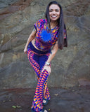 Woman wearing a colorful tie-dye outfit standing against a stone wall.